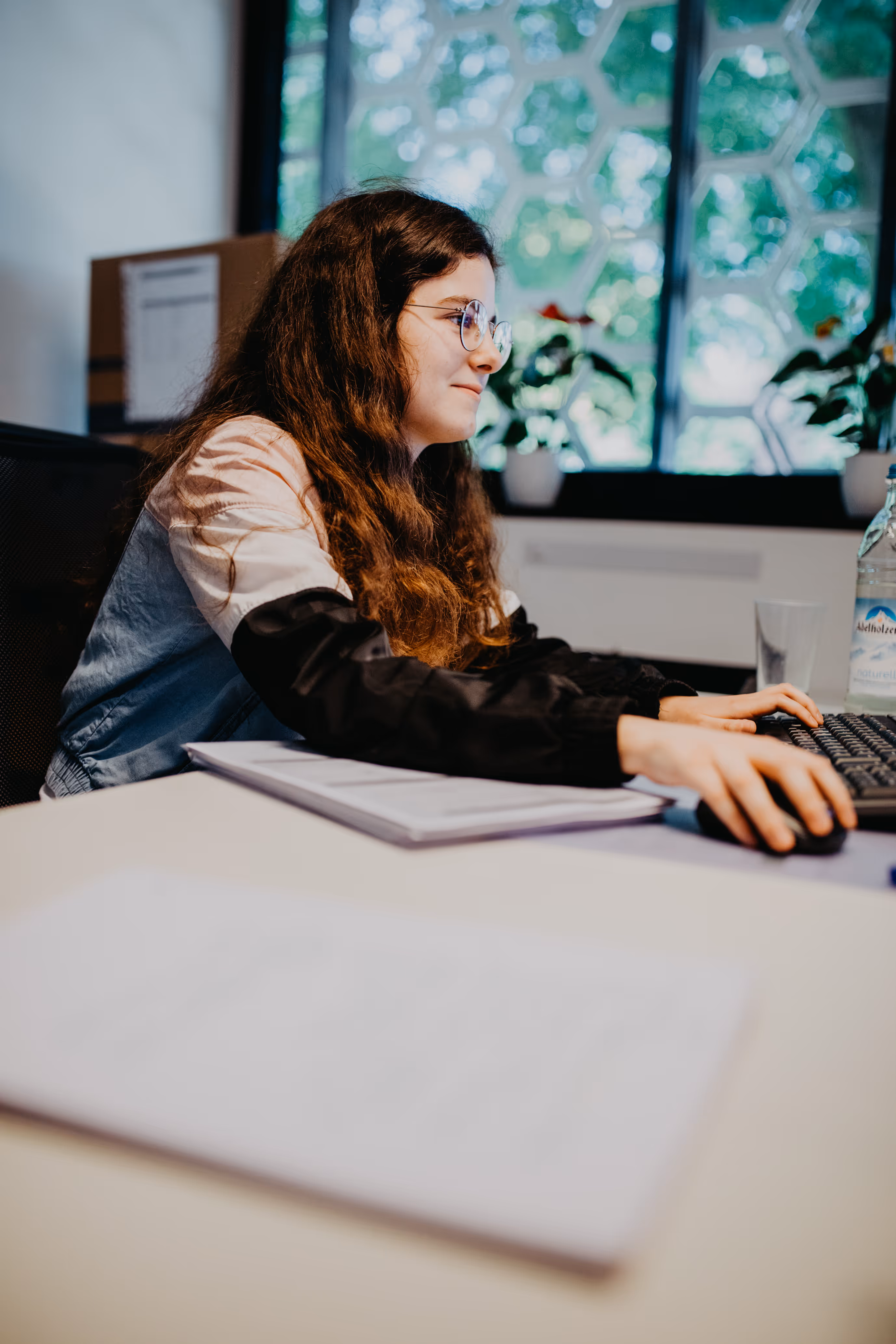 Junge Frau mit Brille arbeitet am Computer in einem hellen Büro mit Pflanzen auf der Fensterbank.