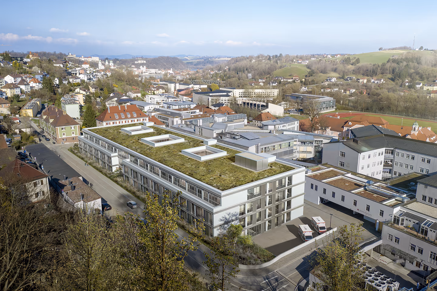 Luftaufnahme eines modernen Gebäudekomplexes mit begrüntem Flachdach in einer Stadtlandschaft mit umliegenden Häusern und grünen Hügeln im Hintergrund.