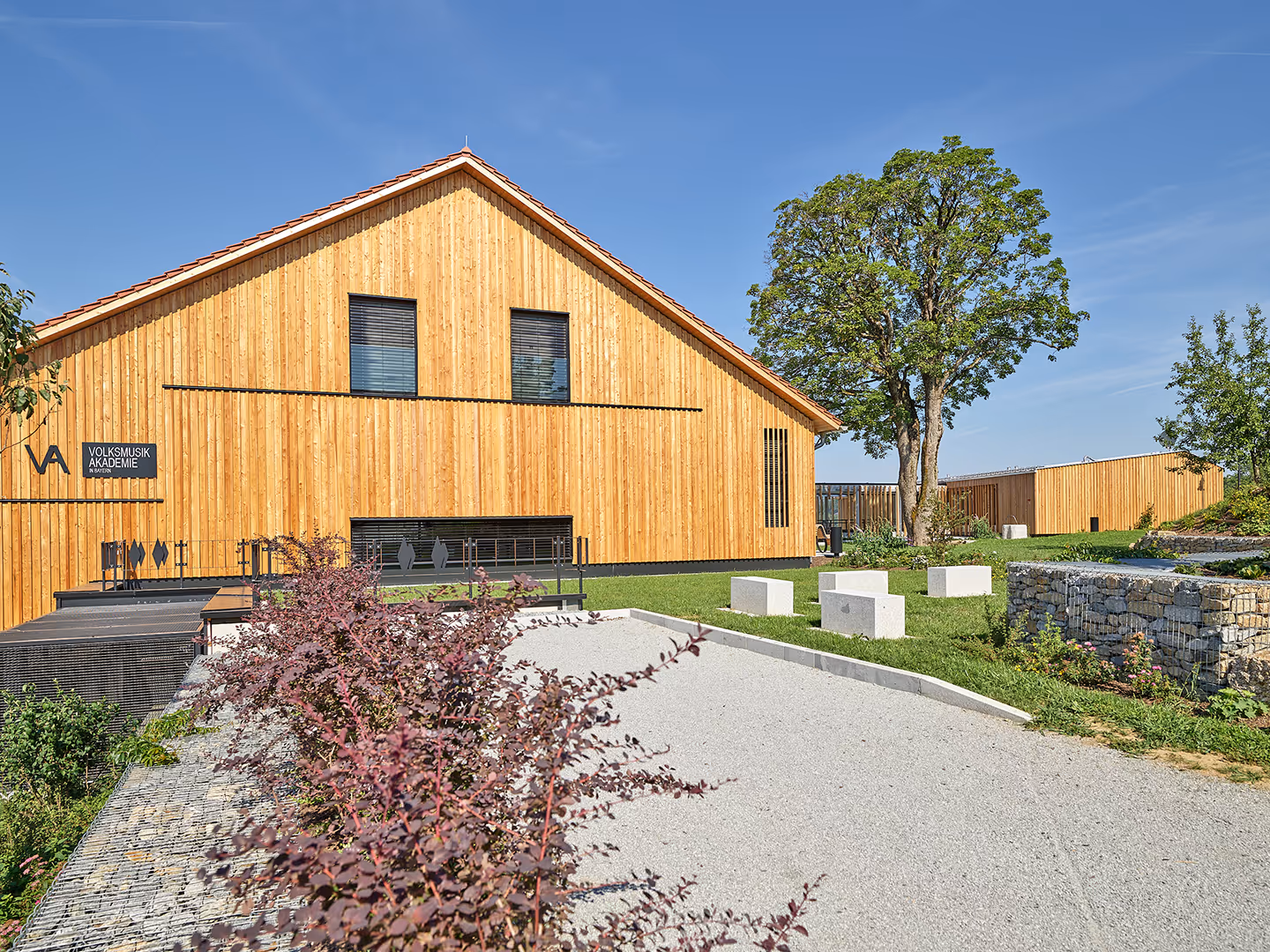 Modernes Holzgebäude der Volksmusik Akademie in Bayern mit grünem Garten, großer Baum und blühenden Pflanzen unter klarem blauem Himmel.