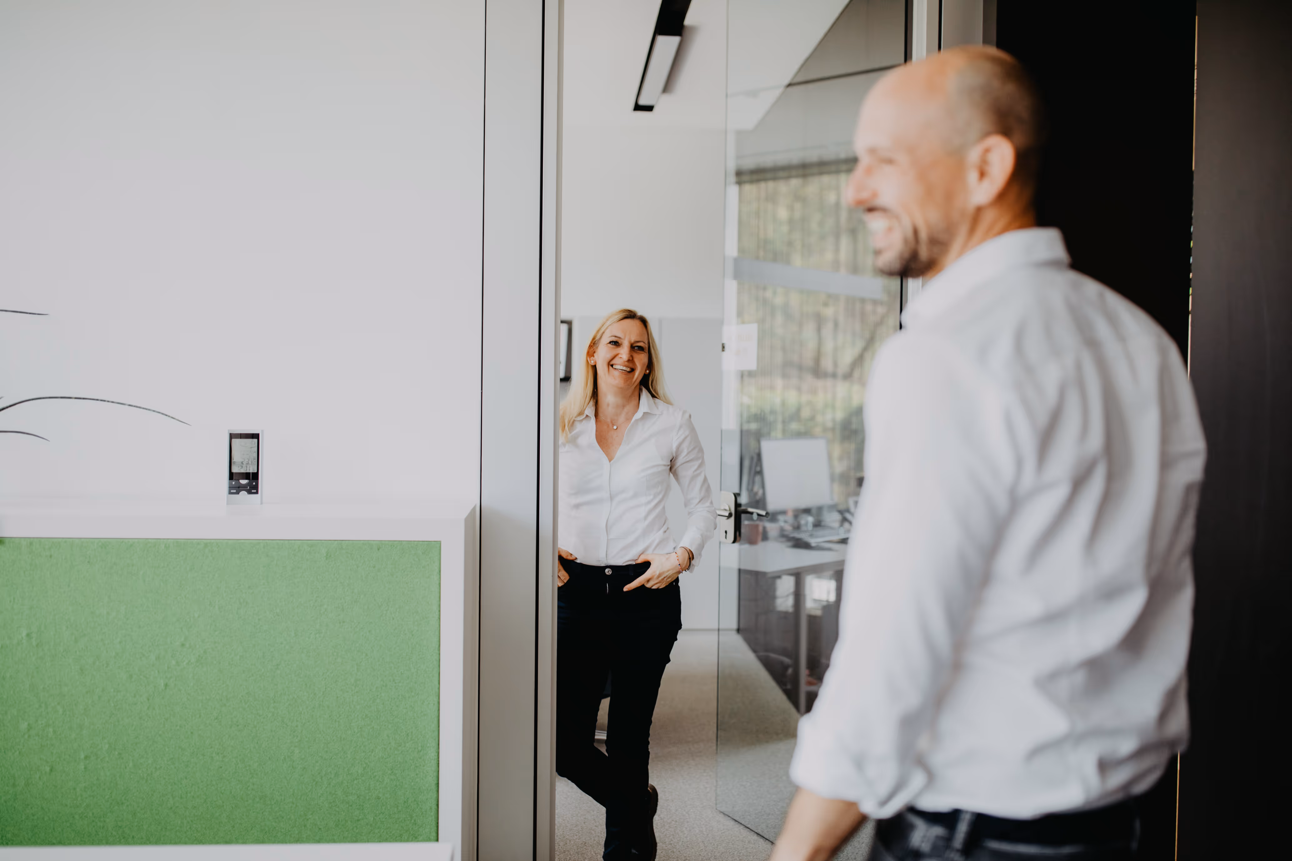 Frau und Mann in einem Büro, die lächelnd gegenüberstehen, die Frau lehnt an einer Türöffnung.