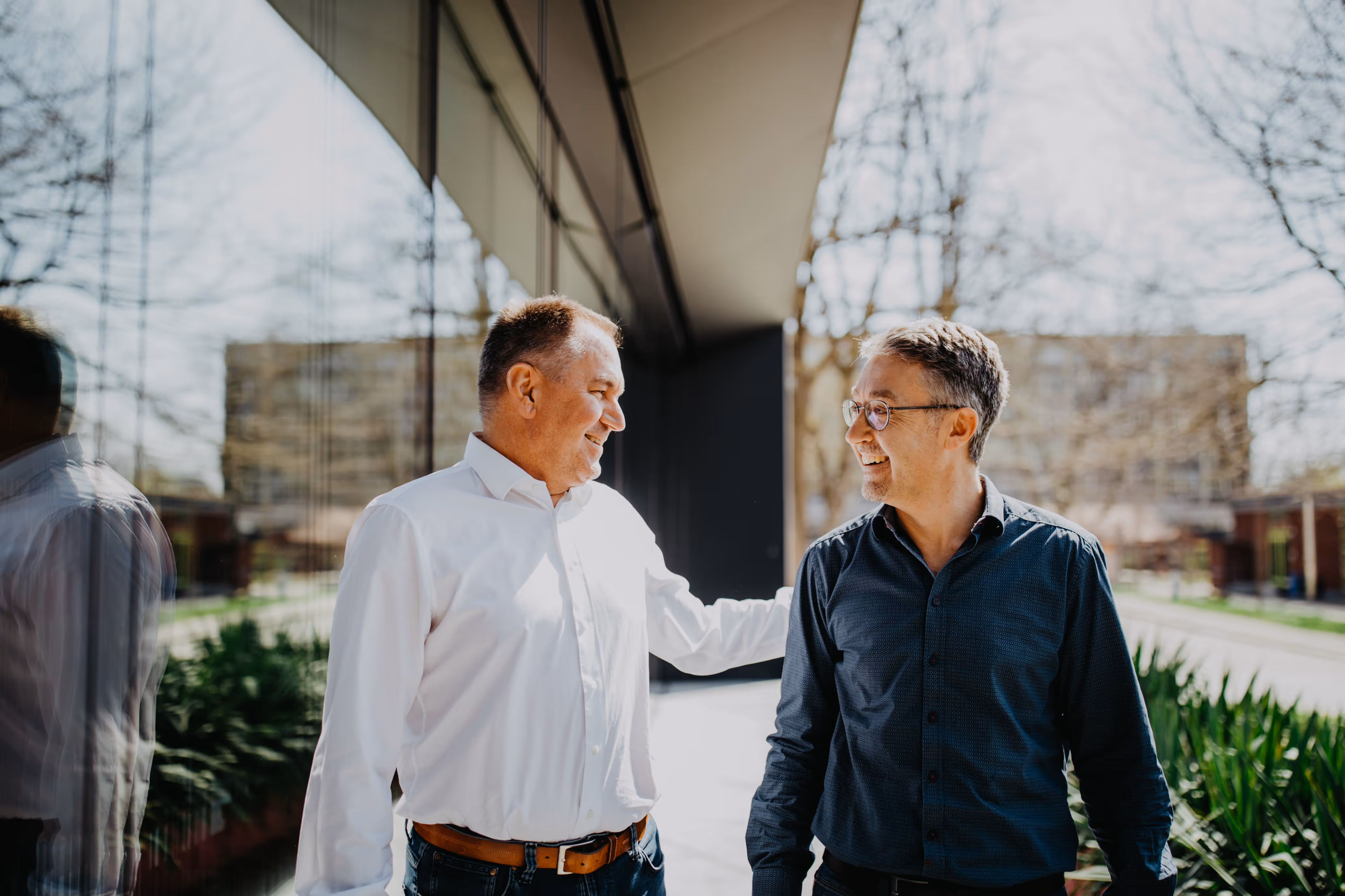 Zwei lächelnde Männer, einer im weißen Hemd, der andere im dunklen Hemd mit Brille, stehen draußen vor einem Gebäude und unterhalten sich.