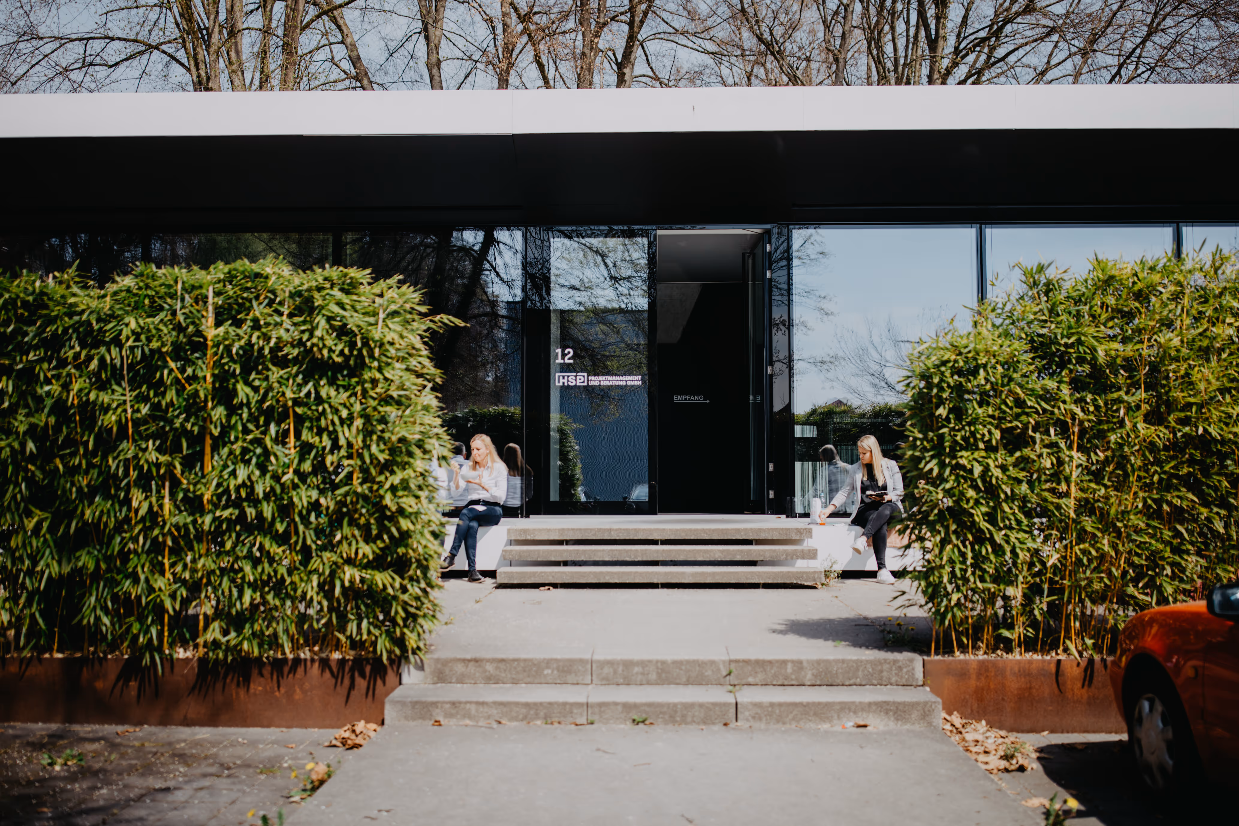 Zwei Frauen sitzen auf beiden Seiten des Eingangs zu einem modernen Bürogebäude mit großen Glasfenstern, umgeben von grünen Büschen.