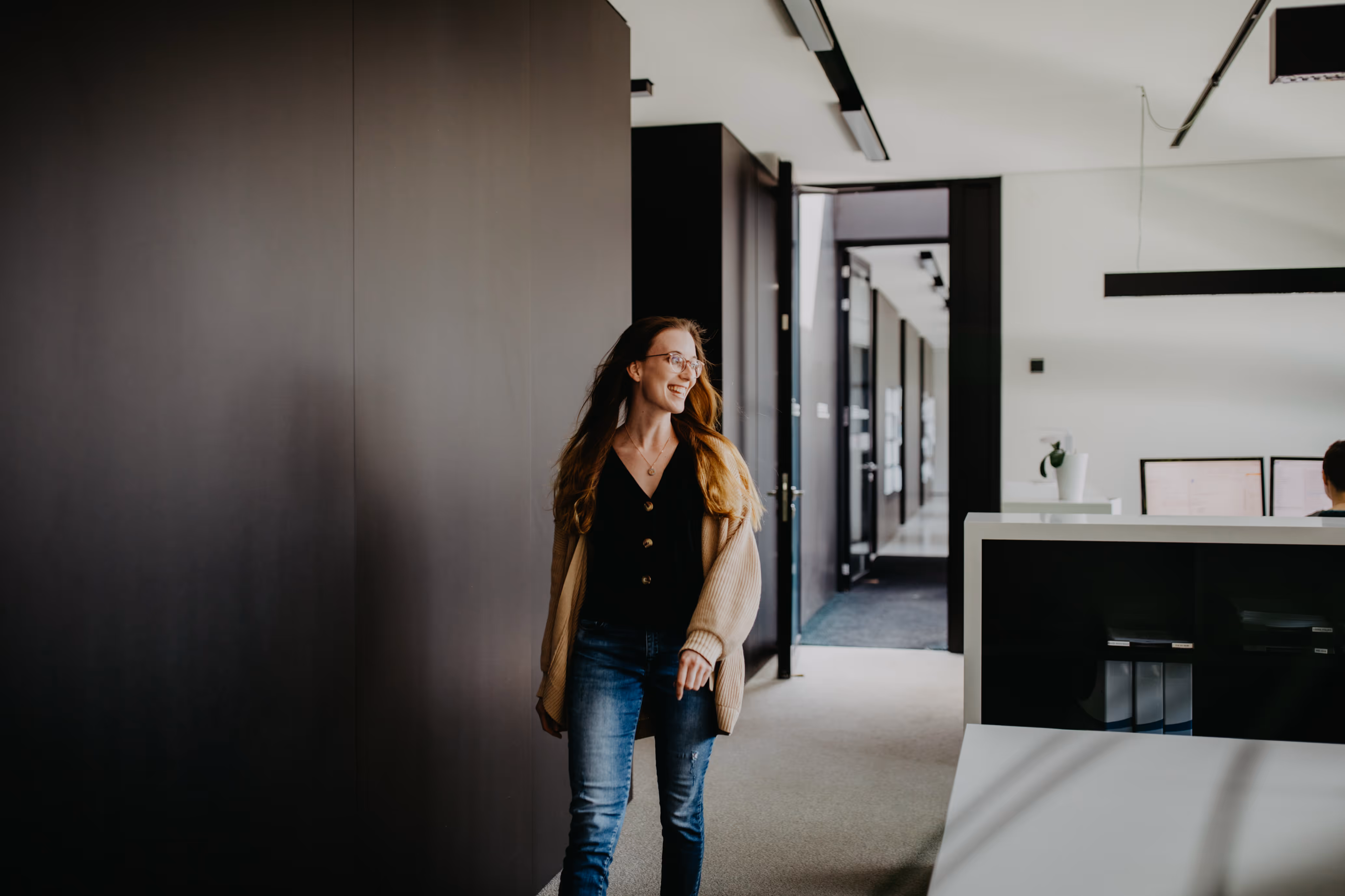 Frau mit braunen Haaren und Brille geht lächelnd in einem modernen Büroflur.