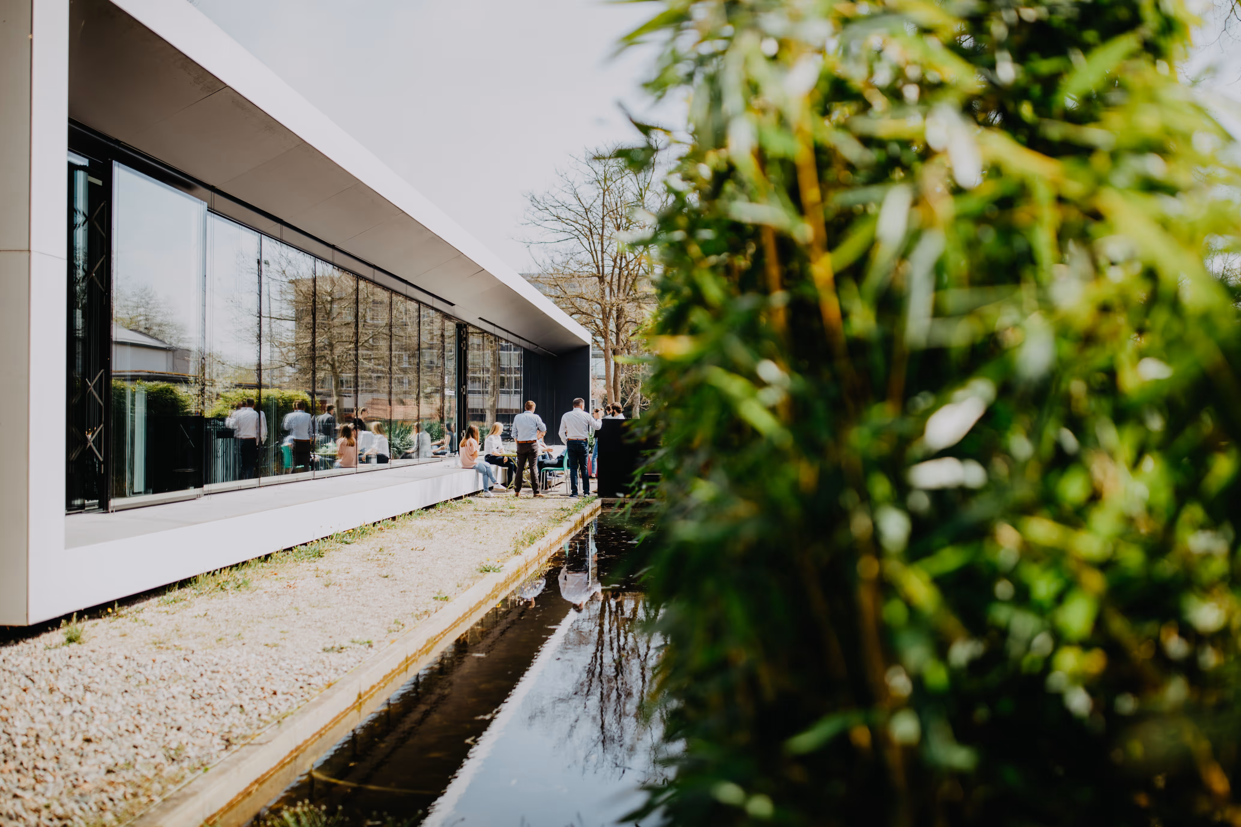 Personen im Freien neben einem modernen Gebäude mit großen Glasfenstern und einem Wasserbecken.