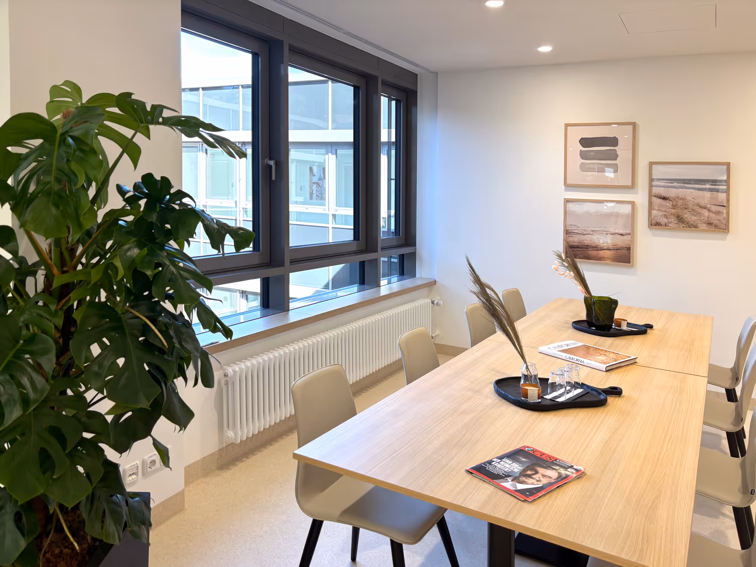 Moderner Besprechungsraum mit großem Holztisch, sechs hellen Stühlen, grüner Pflanze und drei gerahmten Bildern an der weißen Wand.