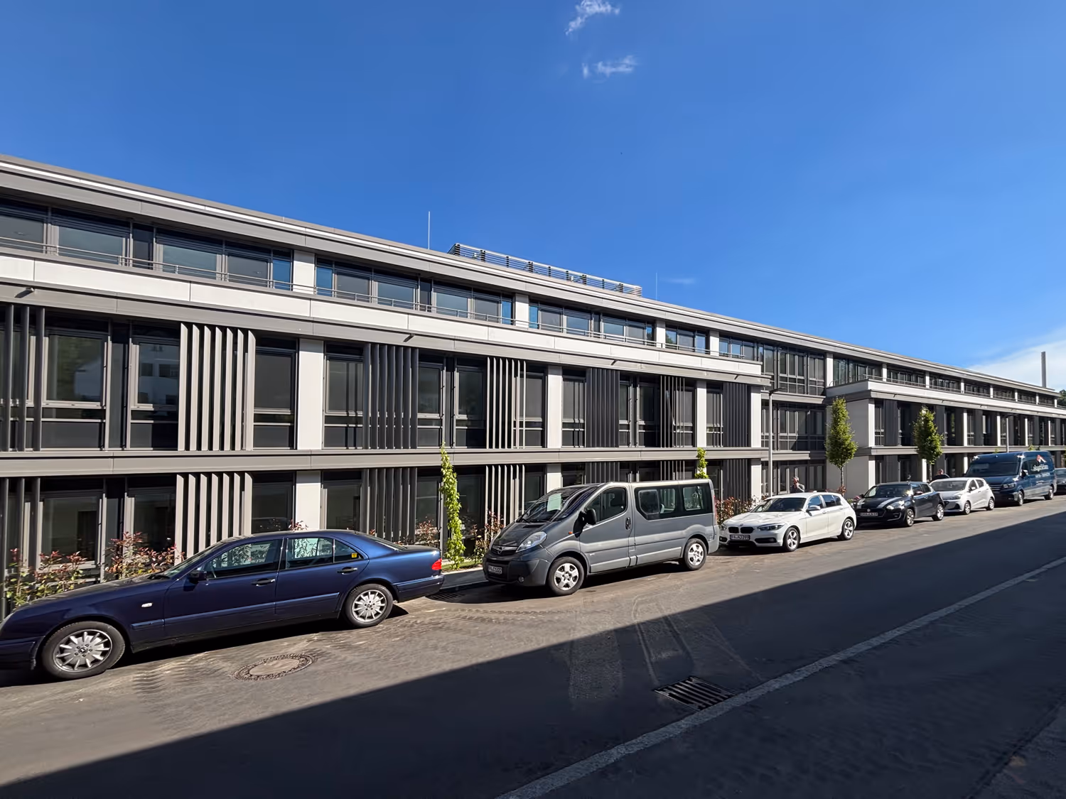 Modern zweistöckiges Klinikum mit vertikalen Lamellen an den Fenstern und mehreren geparkten Autos davor an einem sonnigen Tag.
