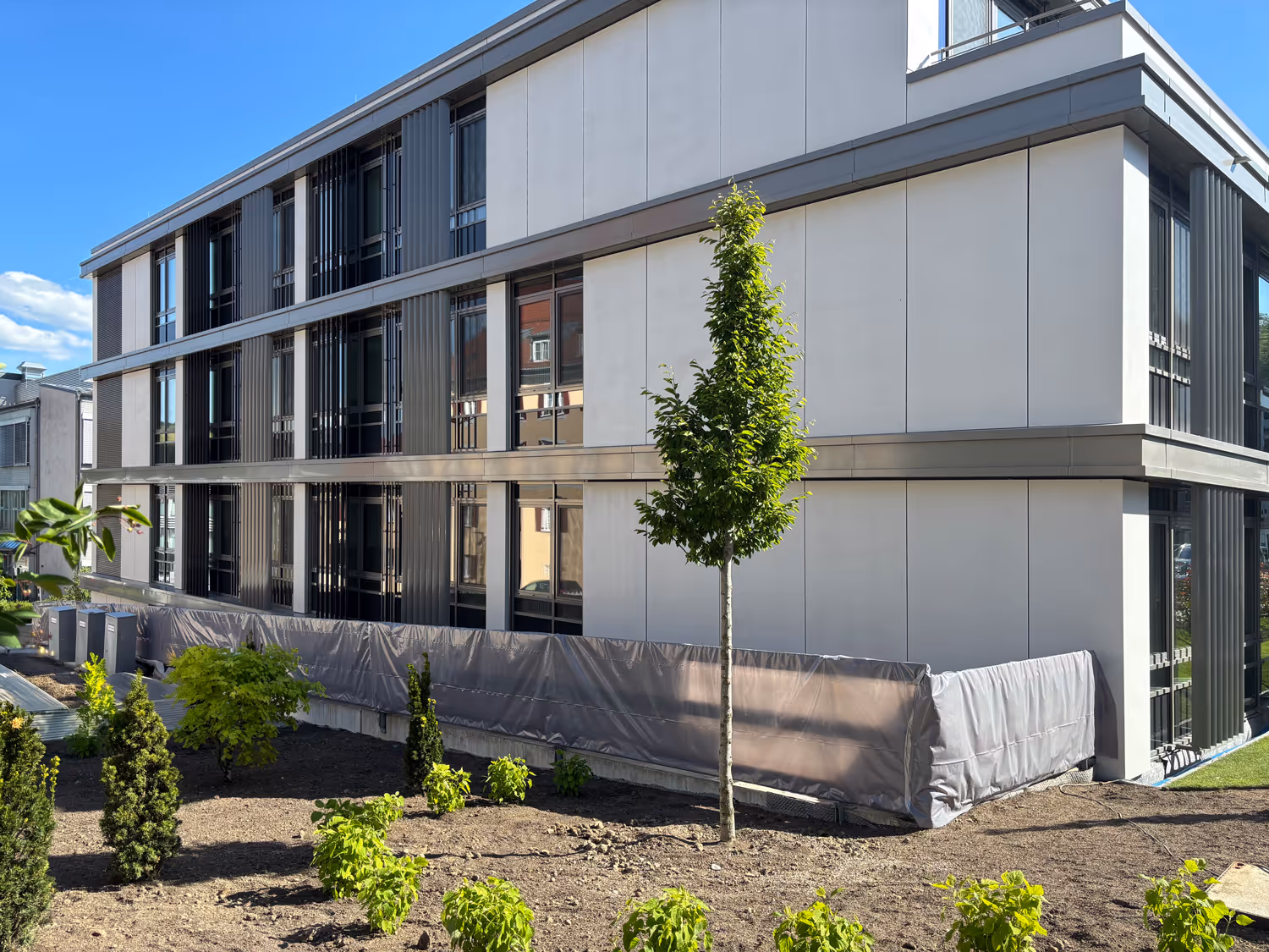 Modernes Klinikums mit hellem Fassadenmaterial und Garten mit jungen Bäumen und Sträuchern unter klarem blauem Himmel.