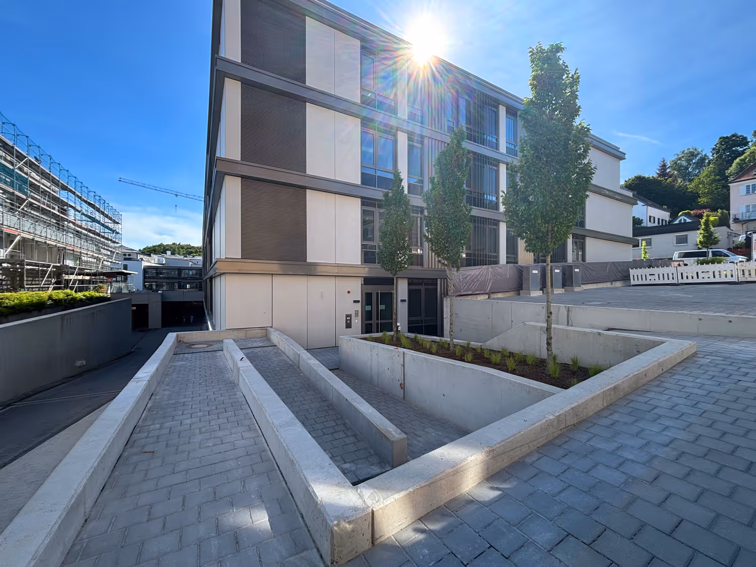 Modernes Klinikum bei Sonnenschein mit gepflastertem Vorplatz, drei Bäumen und einer Rampe mit Betonbegrenzungen.