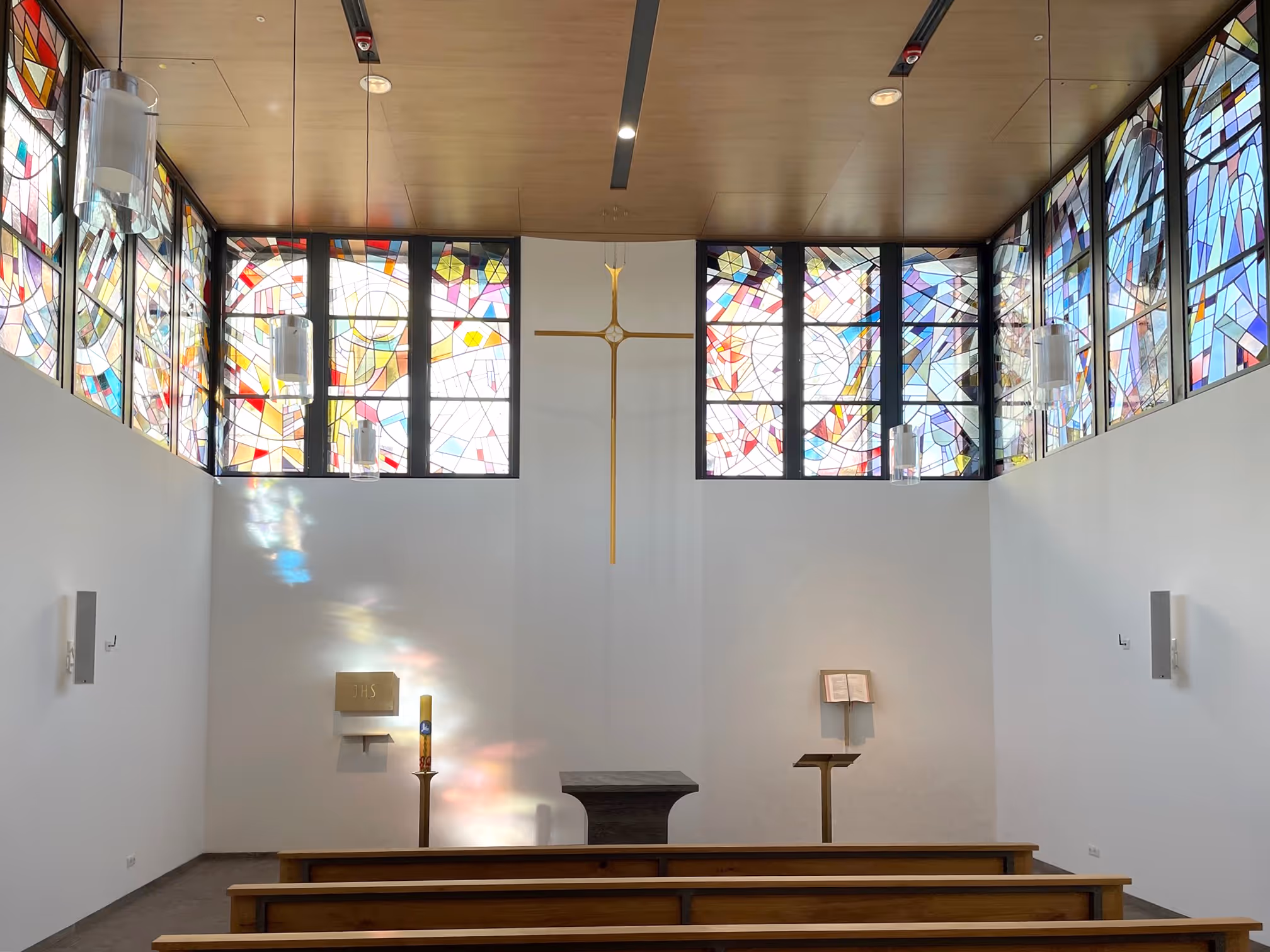 Innenraum einer kleinen Kapelle eines Klinikums mit bunten Glasfenstern, hölzernen Bänken, einem goldenen Kreuz an der Wand, Altar und Kerze.