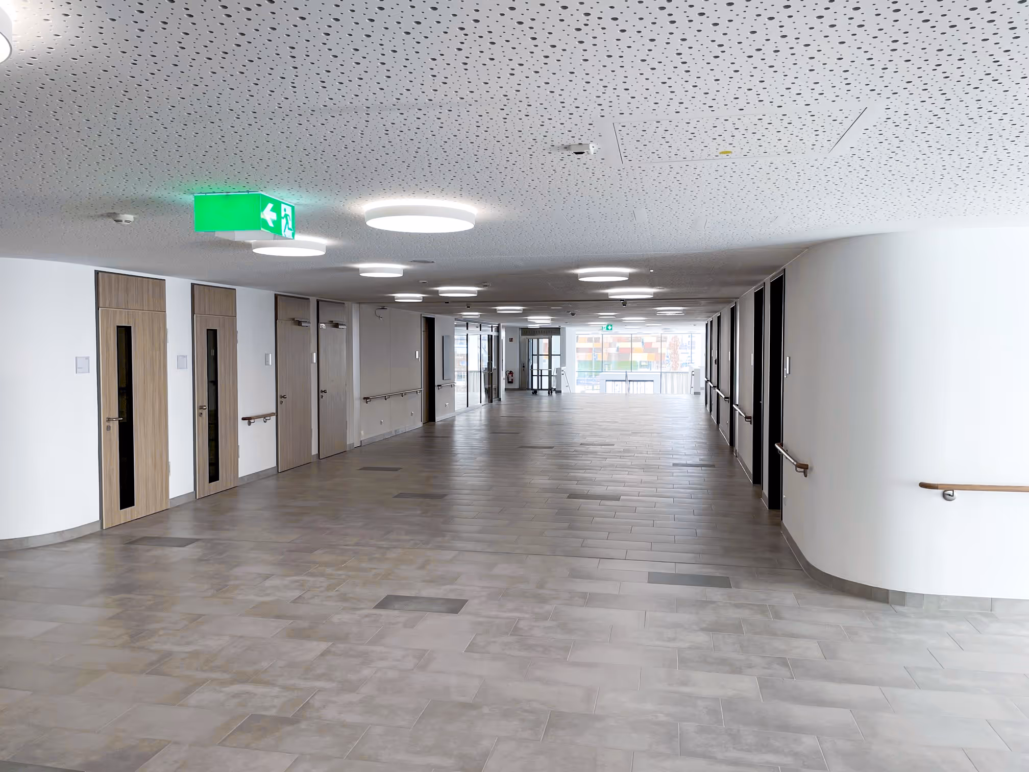 Heller Flur in einem modernen Klinikum mit grauem Fliesenboden, mehreren Türen an den Seiten und Deckenlichtern.