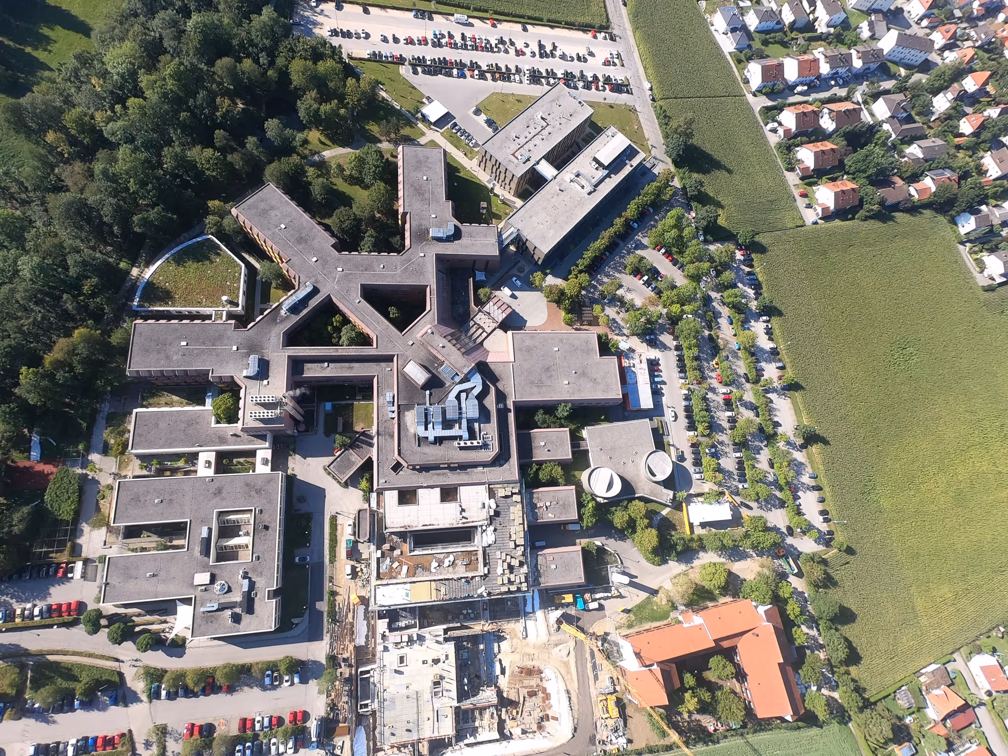 Luftaufnahme eines großen Komplexes von einem Klinikum mit Parkplätzen, grünen Feldern und Wohnhäusern in der Nähe.
