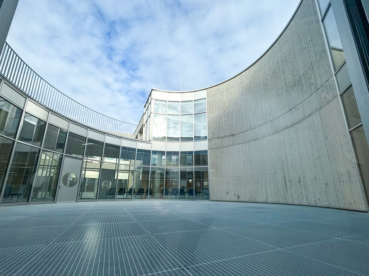 Moderner Innenhof mit gebogener Betonwand, großen Glasfenstern und Metallgitterboden unter bewölktem Himmel.