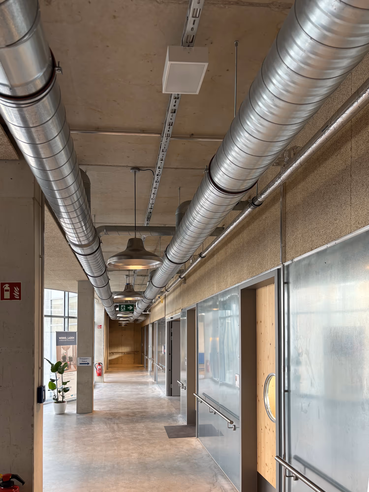 Moderner Gebäudeflur mit sichtbaren Lüftungsrohren an der Decke, mehreren Türen, Handläufen und Notausgangsschildern.