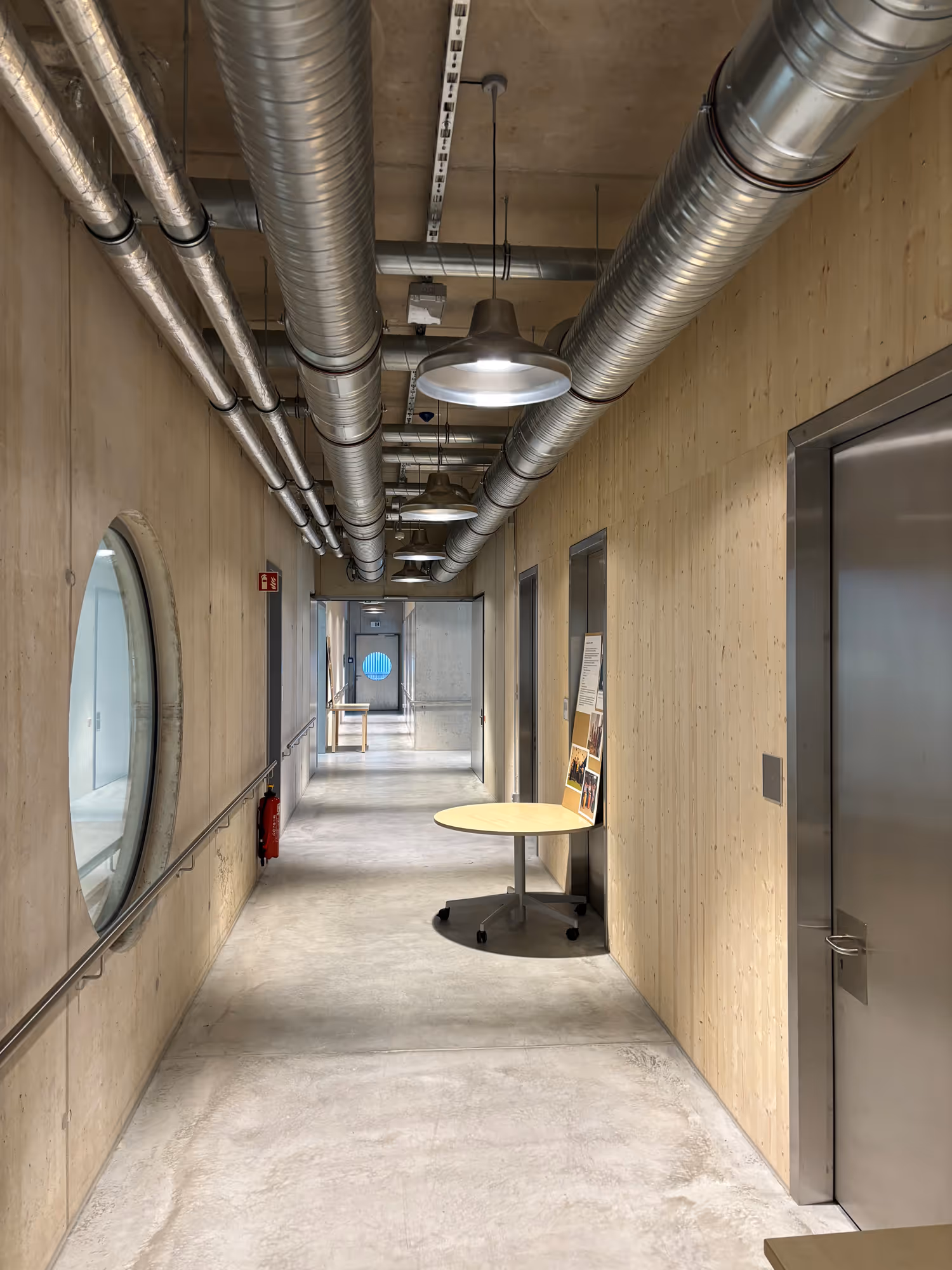 Moderner Flur mit Betonboden, Holzverkleidungen, runden Fenstern, mehreren metallischen Rohren an der Decke und einem runden Tisch auf Rollen.