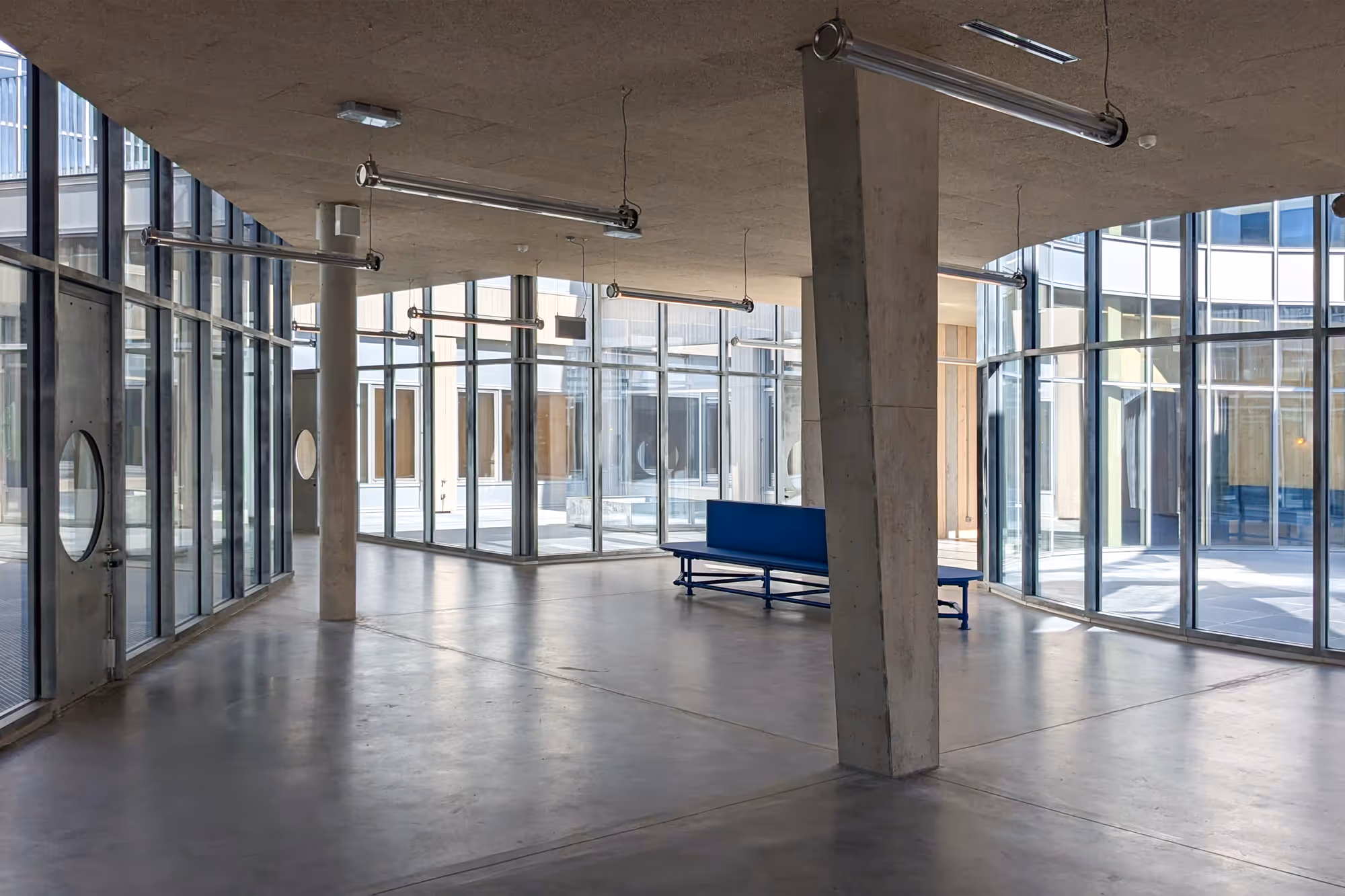Großer, offener Innenraum mit sichtbaren Betonstützen und blauer Bank, umgeben von bodentiefen Glasfenstern.