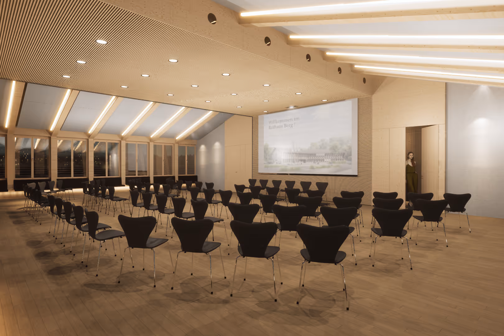 Heller Konferenzraum mit mehreren Reihen schwarzer Stühle, großem Bildschirm mit Text ‚Willkommen im Rathaus Berg!‘ und einer Frau, die in der Tür steht.