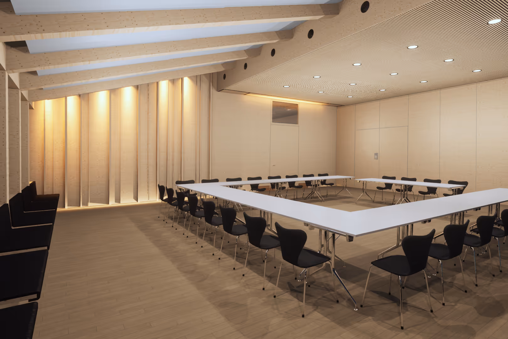 Moderner Raum mit U-förmigen weißen Tischen und schwarzen Stühlen, Holzverkleidung und Beleuchtung an der Decke.