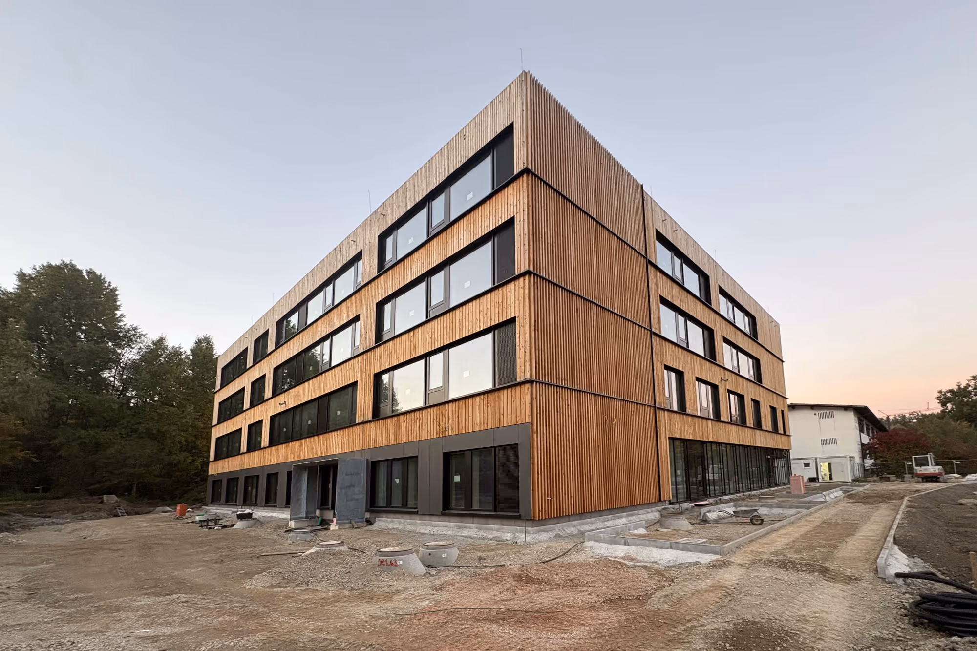 Modernes mehrstöckiges Gebäude mit Holzfassade im Bau, umgeben von unbefestigtem Boden und Bäumen im Hintergrund.