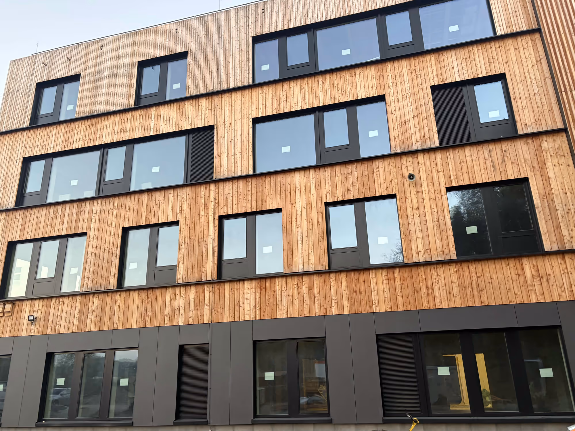 Moderne Gebäudehülle mit Holzverkleidung und schwarzen Fensterrahmen, vierstöckig.