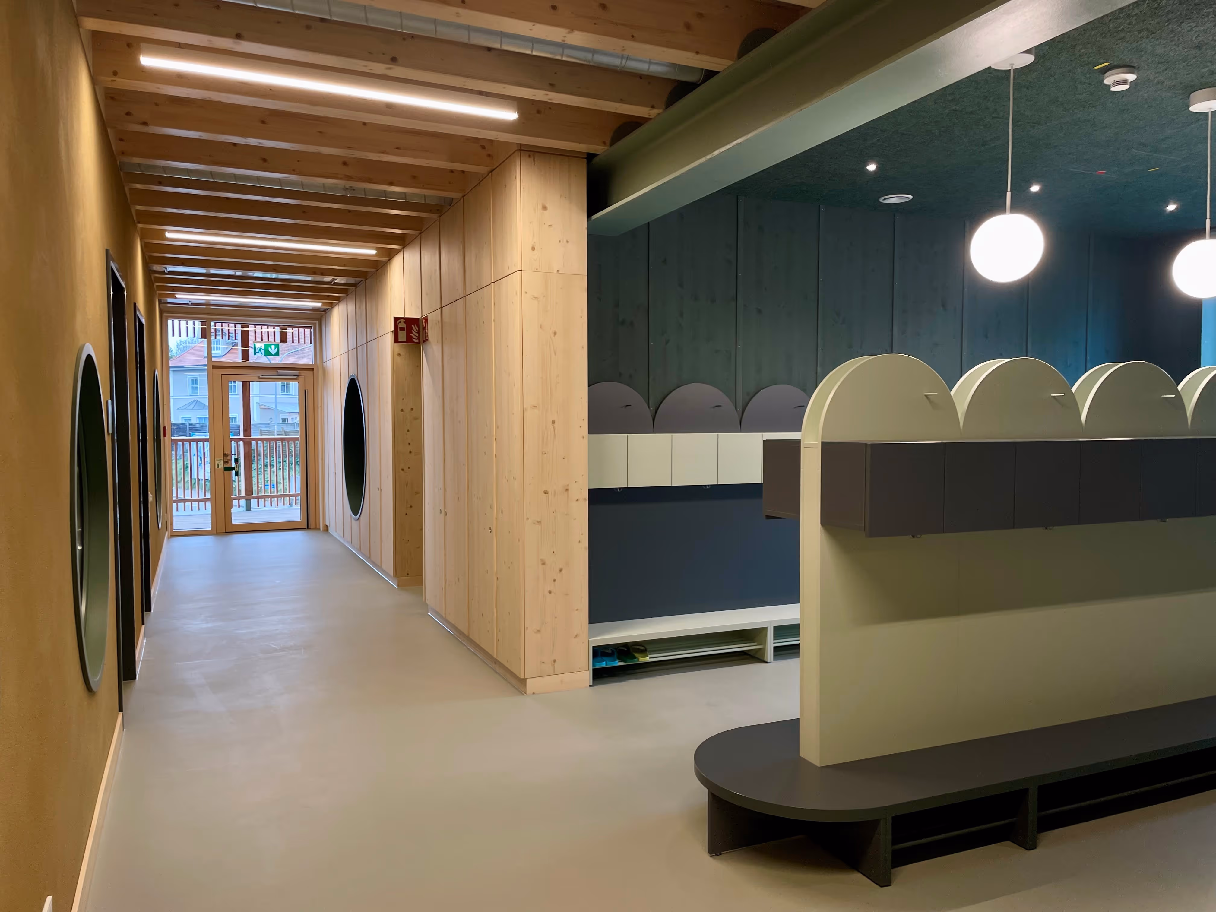 Moderner Flur in einem Gebäude mit Holzwänden, rundem Fenster und Garderobenbereich mit Hockern unter Hängeleuchten.