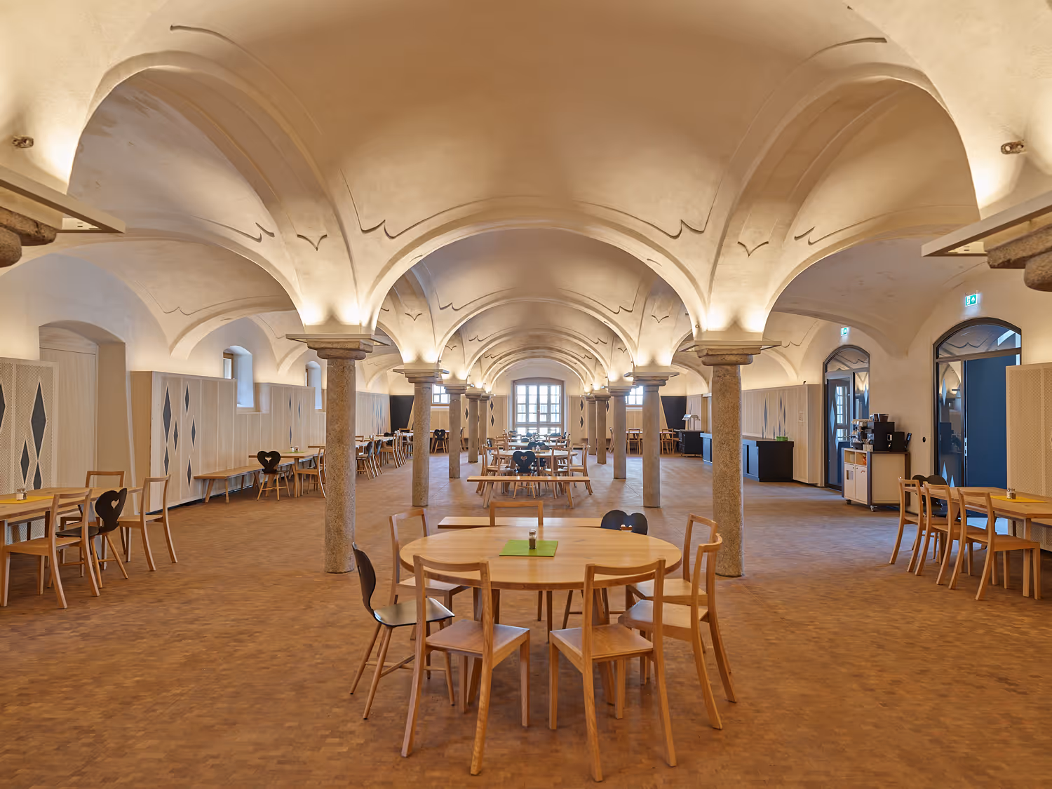 Großer Speisesaal mit hölzernen Tischen und Stühlen, gewölbter Decke und mehreren Steinsäulen.