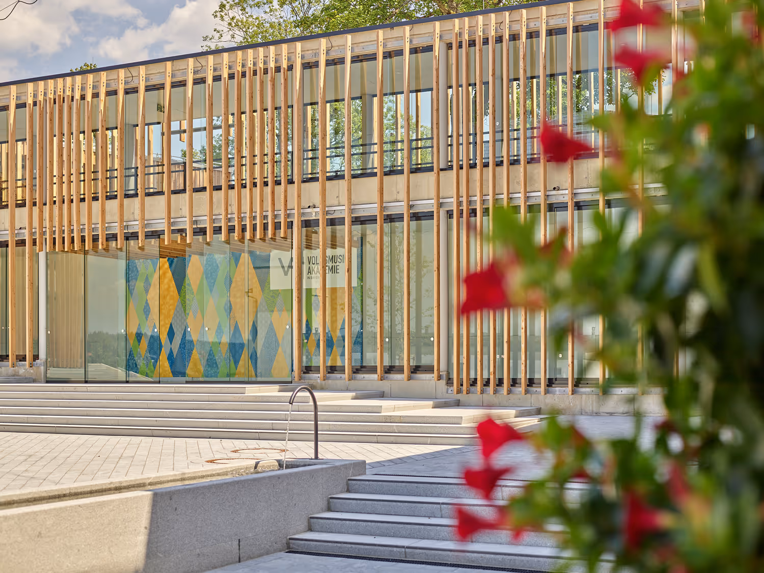 Moderne Gebäudefassade mit vertikalen Holzlatten, großen Glasfenstern, Stufen und verschwommenen roten Blumen im Vordergrund.