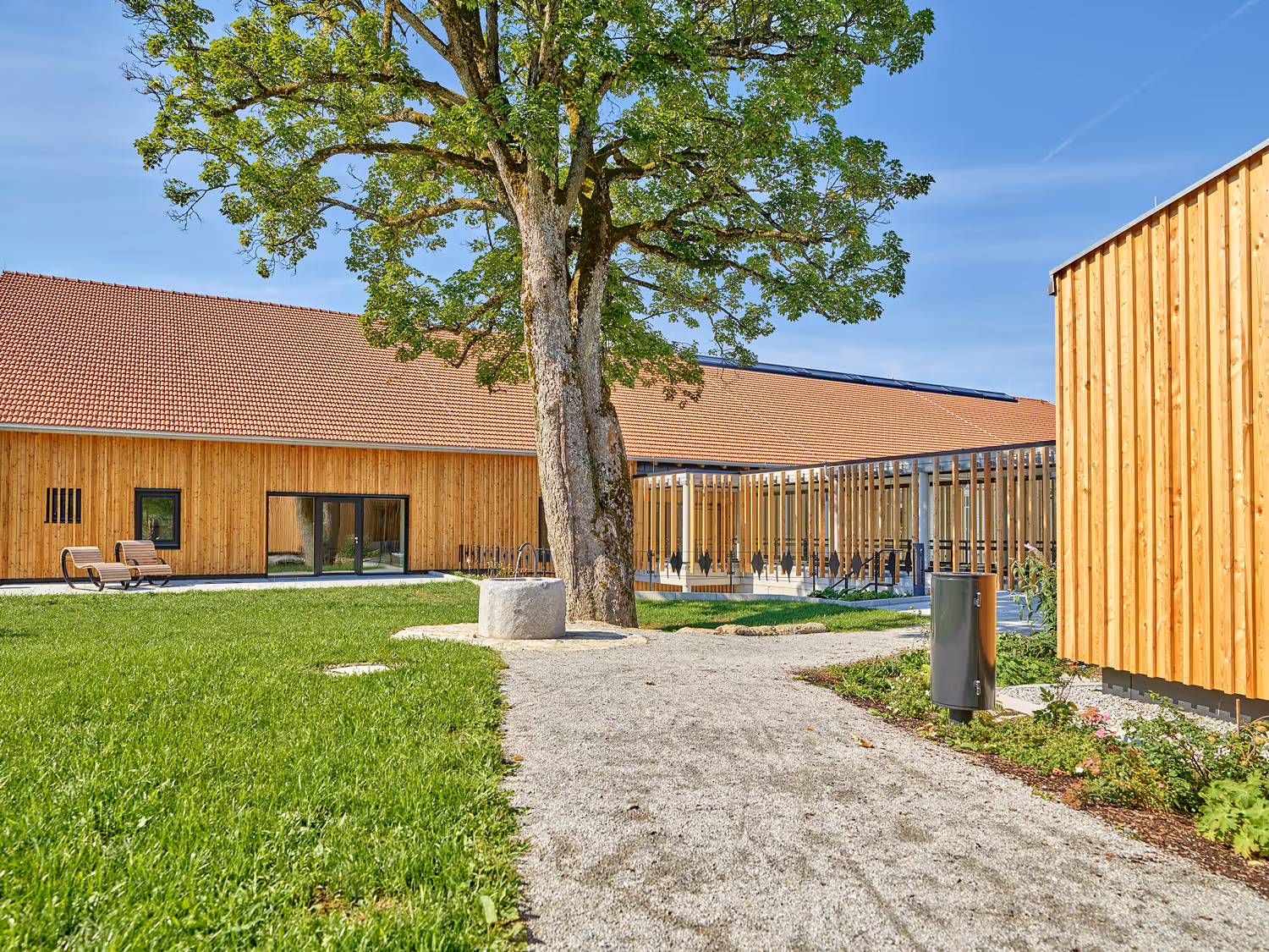 Gepflegter Innenhof mit großem Baum, Sitzbank, Holzgebäuden mit Ziegeldächern und einem Kiesweg.
