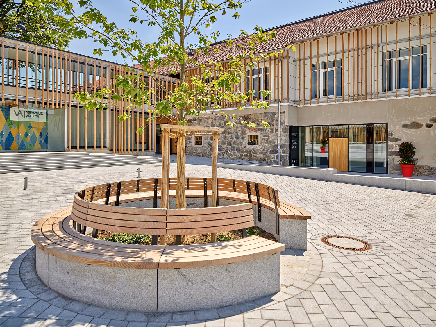 Moderner Innenhof mit rundem Holzbankkreis um einen jungen Baum vor einem Gebäude mit Holzvertäfelung und großen Fenstern.