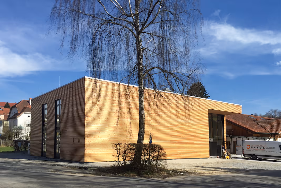 Modernes Gebäude mit Holzverkleidung, daneben ein Baum ohne Blätter und ein Lieferwagen bei sonnigem Himmel.