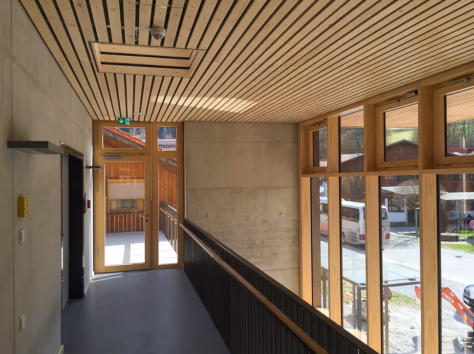 Flur mit großem Fenster und Holztür, Holzbalkendecke und Betonwand, Blick auf eine Straße.