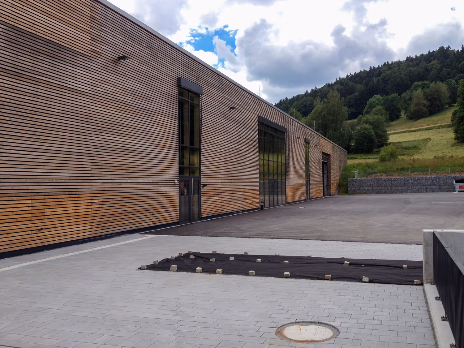 Moderne Holzwand eines Gebäudes mit hohen Fenstern und freier gepflasterter Fläche im Vordergrund, umgeben von grünen Hügeln.