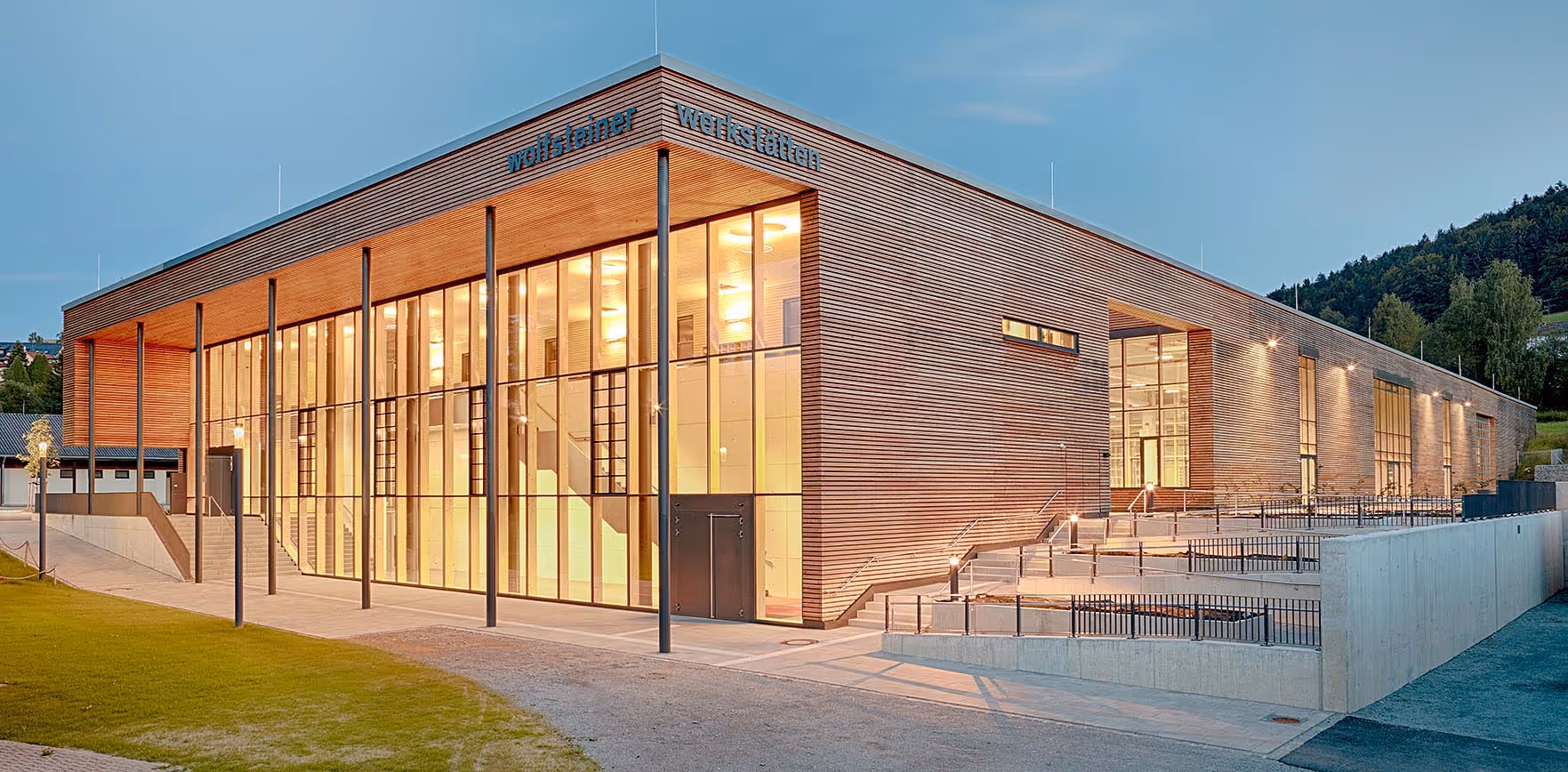 Modernes, langes Gebäude mit großflächigen Glasfenstern und Holzfassade bei Dämmerung.