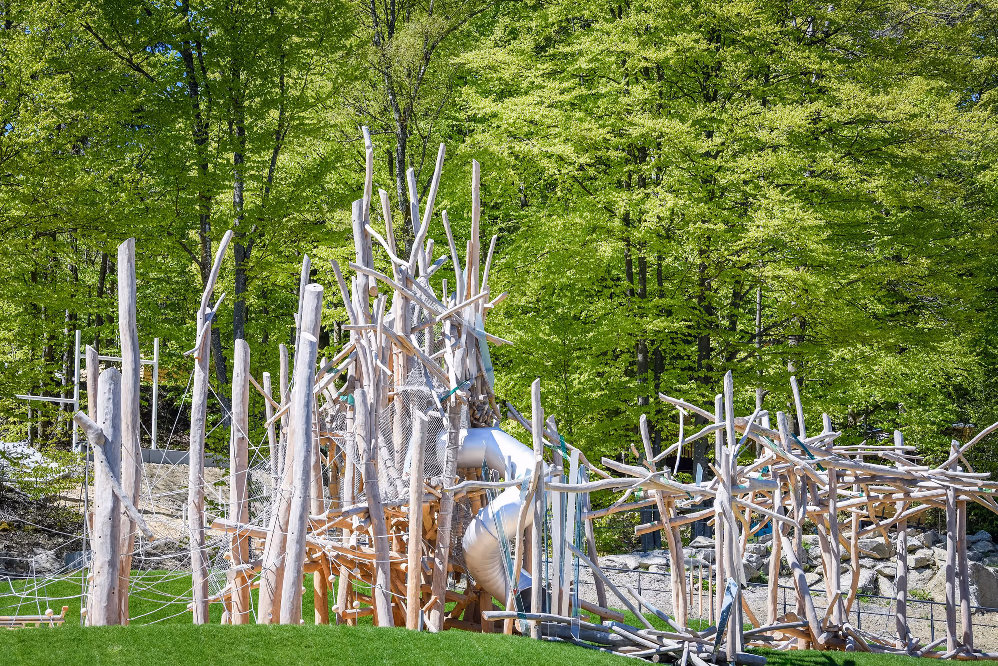 Abenteuerspielplatz aus Holz mit Kletternetzen und Rutsche vor grünem Wald.