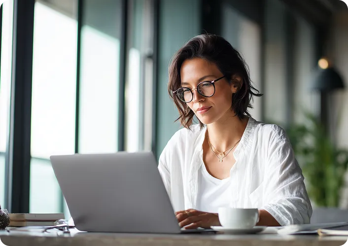 Woman in glasses working on a laptop beside a large office window, with a coffee cup on the desk