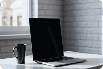Black laptop and coffee mug on white desk near brick wall window