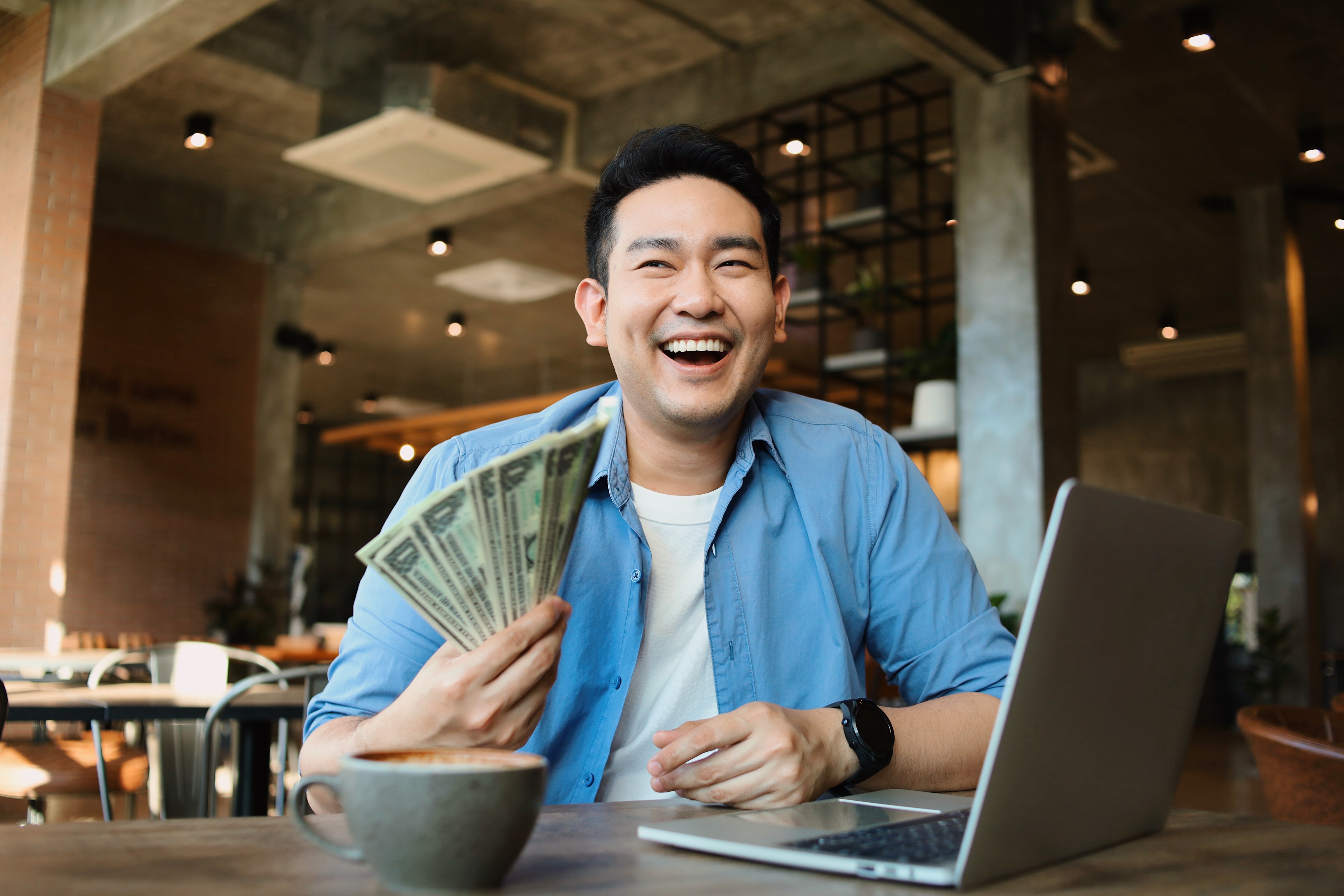 Excited man holding cash, smiling at laptop in coffee shop