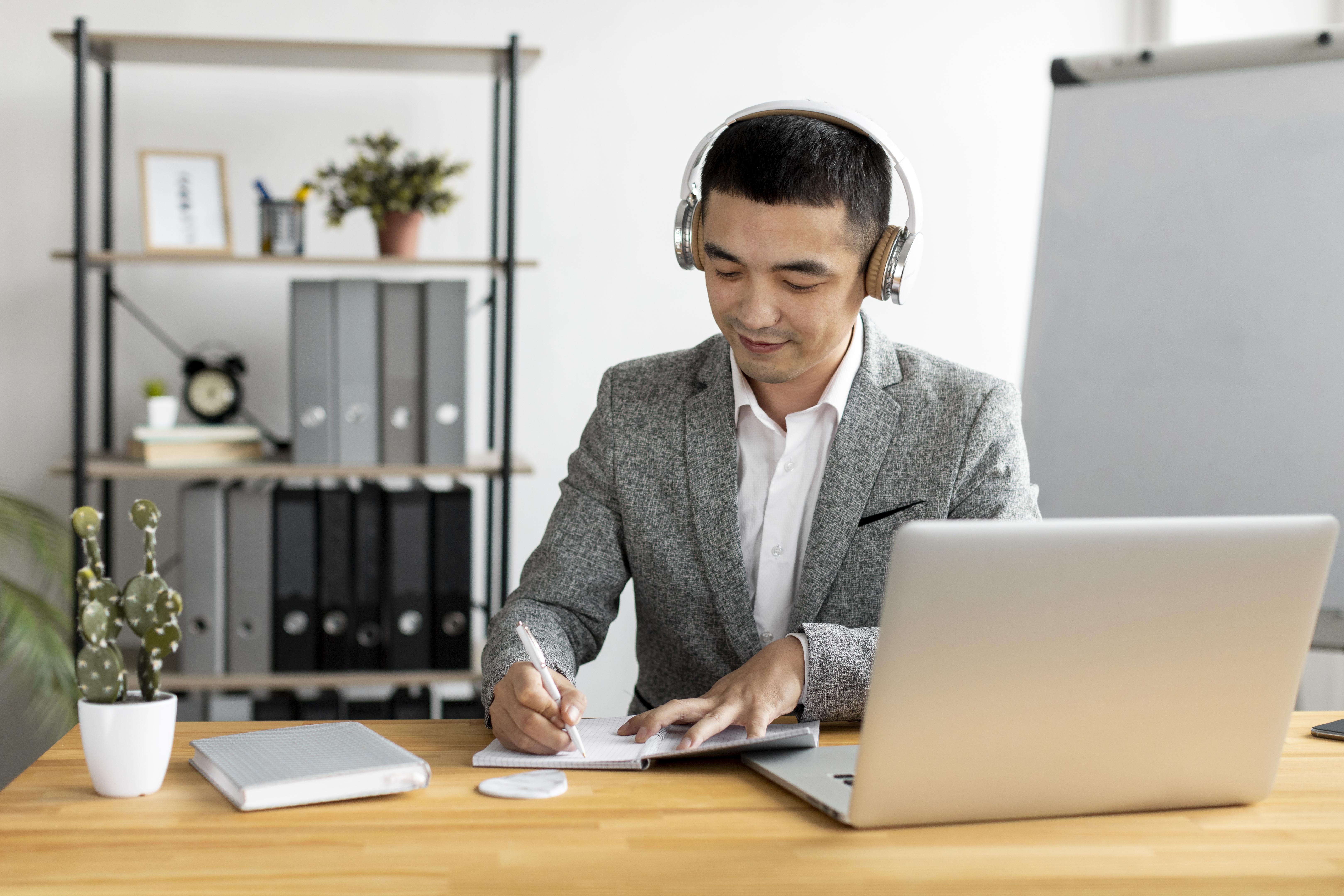 Professional working at desk with headphones, taking notes on laptop