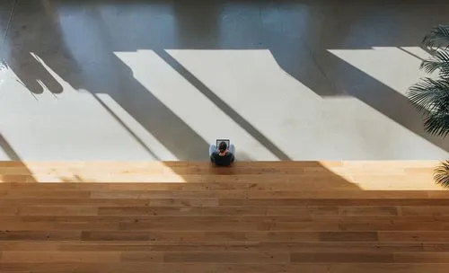 Person working alone on a laptop while sitting on wide wooden steps inside a modern open building with sunlight and shadows
