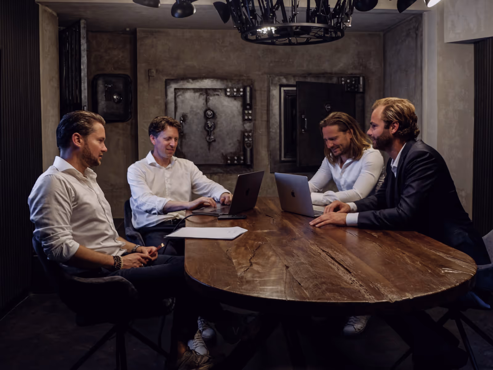 Four men in business attire seated around a wooden table in a dimly lit room, two of them working on laptops.
