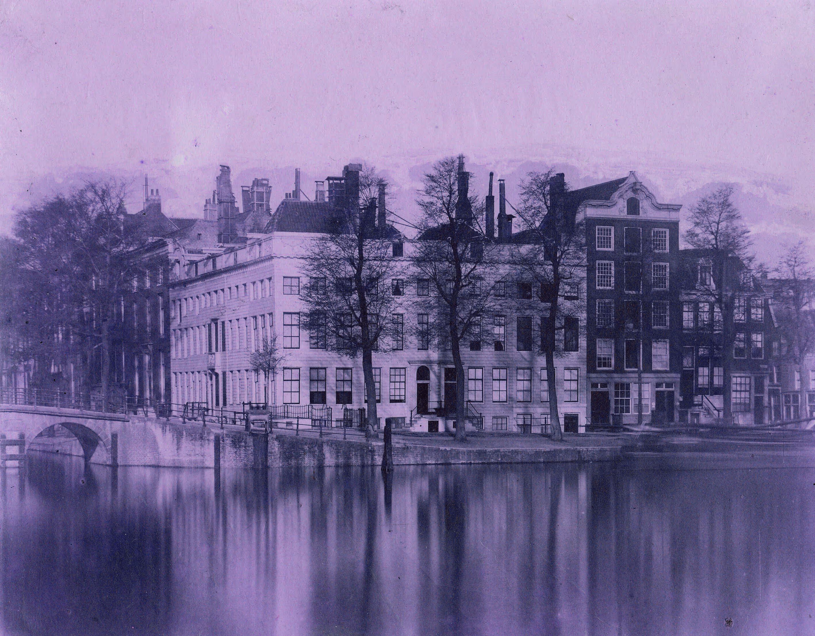 Historic buildings along a canal with leafless trees and a stone bridge reflected in still water.
