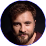Portrait of a man with brown hair and beard looking directly at the camera against a dark background.