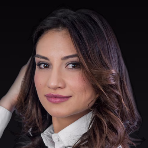 Close-up portrait of a woman with wavy brown hair, wearing a white shirt and looking confidently at the camera against a black background.
