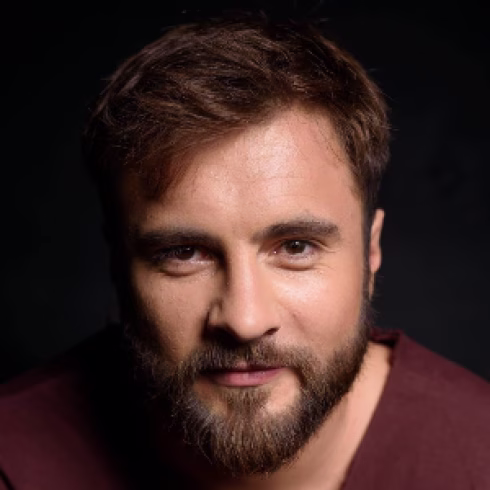 Close-up of a young man with short brown hair and beard against a dark background.