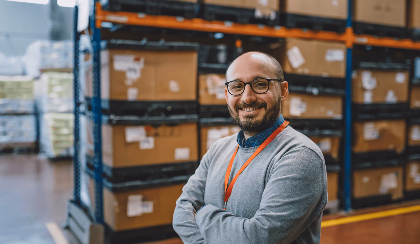 Lächelnder Mann mit Brille und rotem Ausweis vor gestapelten Kartons im Lager.