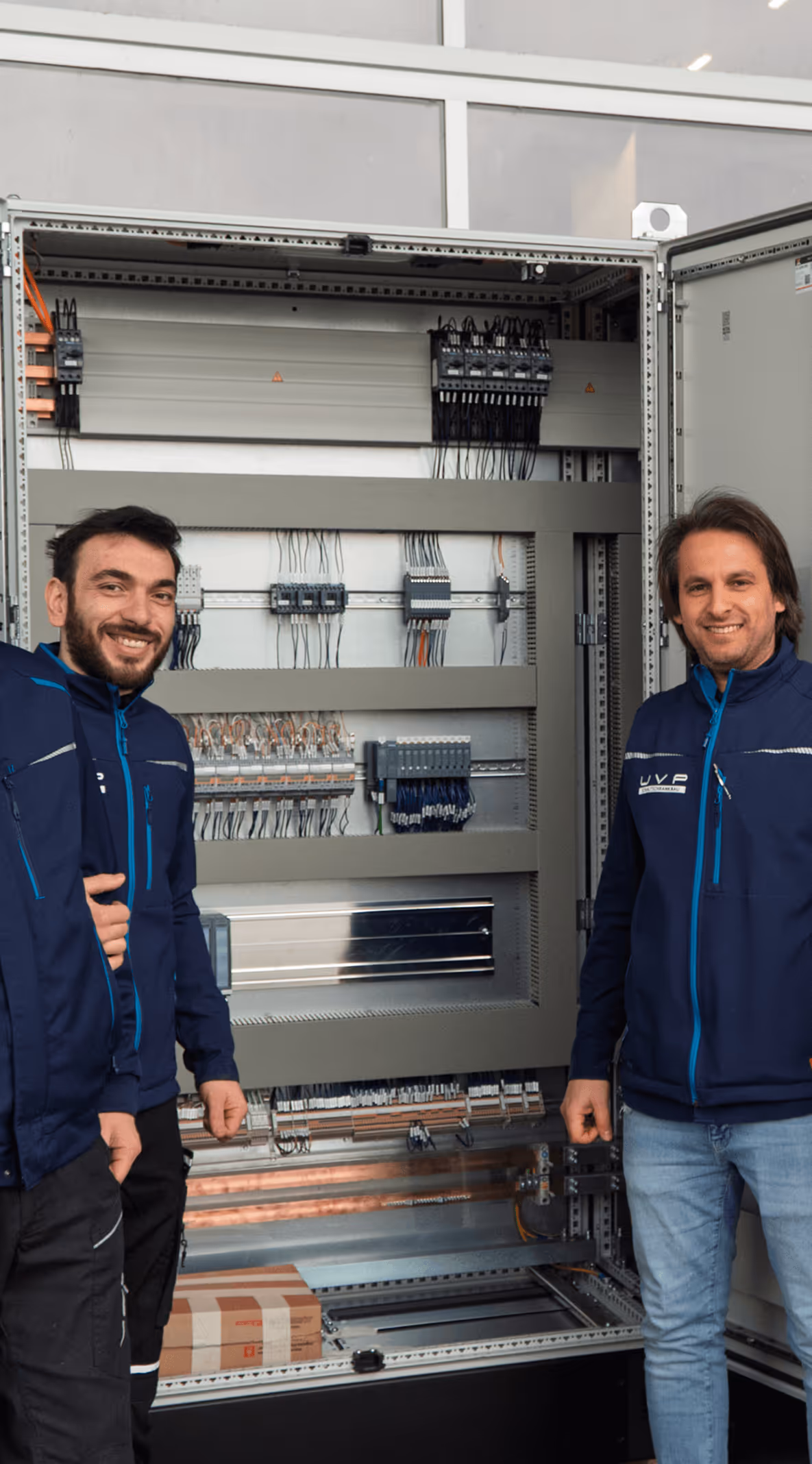 Zwei Männer in blauen Jacken stehen vor einem geöffneten Schaltschrank mit vielen Kabeln und elektrischen Komponenten.
