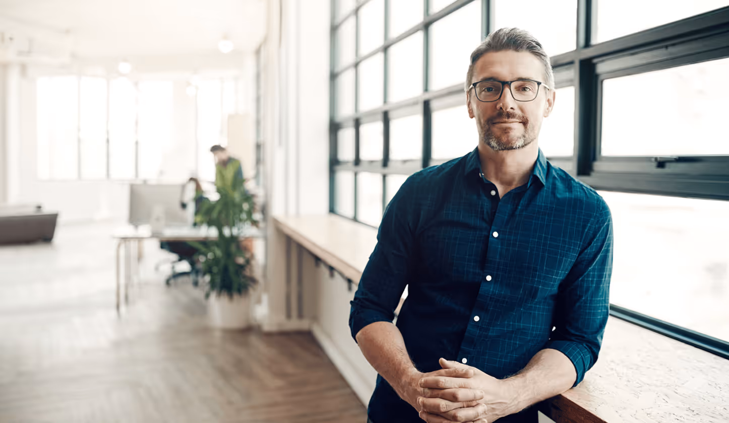 Mann mit Brille lehnt an Fensterbank in modernem Büro, im Hintergrund Kollegen an Schreibtischen.