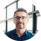 Mann mit Brille und Bart in modernem Büro, Fenster im Hintergrund.