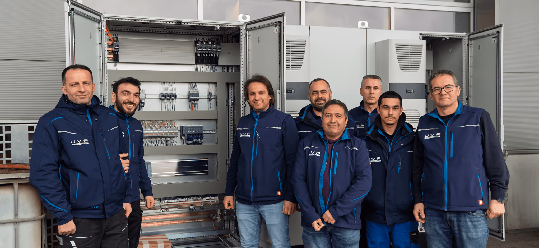 Gruppe von acht Technikern in blauer Arbeitskleidung vor geöffneten Schalttafeln.