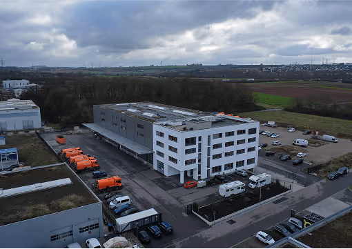 Moderne Lagerhalle mit weißen und grauen Fassaden, parkende orangefarbene LKWs und Autos auf einem bewölkten Tag.