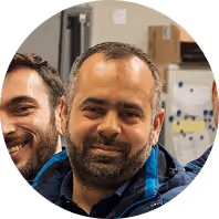 Lächelnder Mann mit Bart in blauer Jacke vor Bürohintergrund.